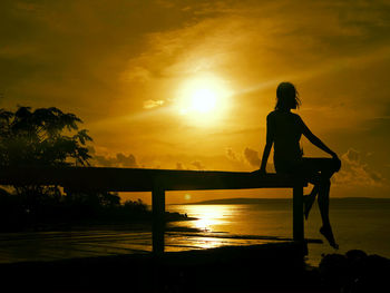 Silhouette woman sitting on railing against sea during sunset