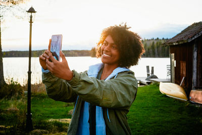 Smiling young woman taking selfie with smart phone while standing against lake during sunset