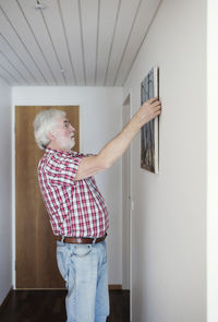 Side view of senior man hanging painting on white wall at home