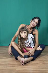 Portrait of mother and daughter sitting on wooden floor against dark green background. 