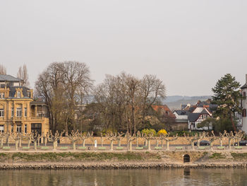 Trees and houses on field by lake against clear sky