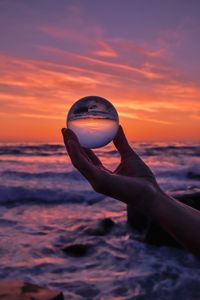 Close-up of hand holding crystal ball against sea during sunset