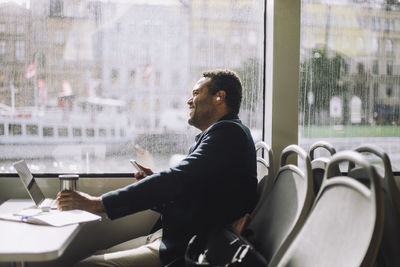 Side view of male freelancer sitting with smart phone and insulated drink container at table in ferry