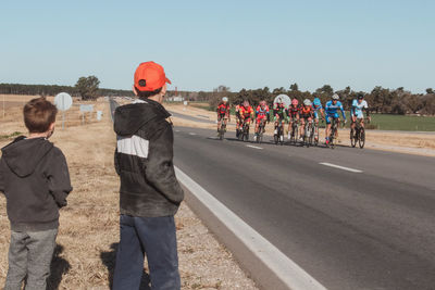 Side view of boys looking at cyclists riding bicycles on road against clear sky