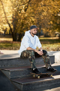 Full length of man sitting by skateboard outdoors