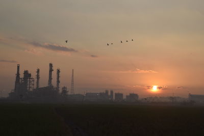 Scenic view of field by industrial buildings against sky during sunset
