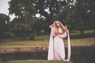 Seductive woman wearing dress standing at lakeshore in forest