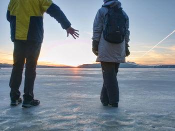 Tourist couple is spending time outdoors on a frozen lake, watching rising sun at horizon.