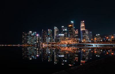 Illuminated modern buildings by river against sky at night