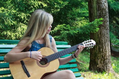 Young woman playing guitar while sitting on bench at park