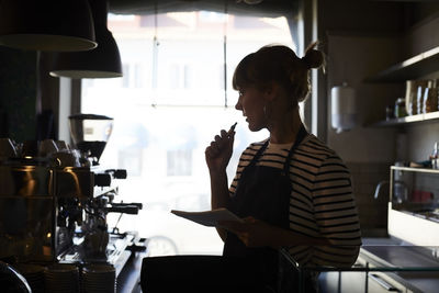 Thoughtful barista holding note pad while looking at coffee maker in cafe