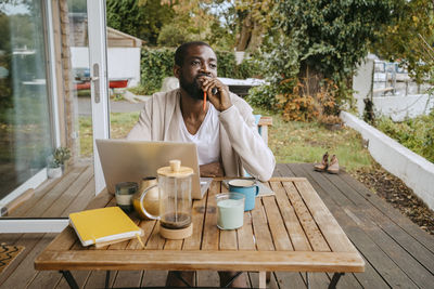 Thoughtful man with laptop sitting at table on porch