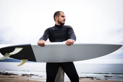 Man holding surfboard standing at beach against sky