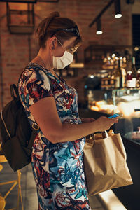 Mature woman wearing mask using mobile phone standing in cafe
