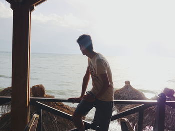 Man sitting on railing while looking at sea during sunny day