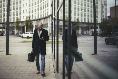 Full length of businessman using mobile phone while walking on sidewalk in city