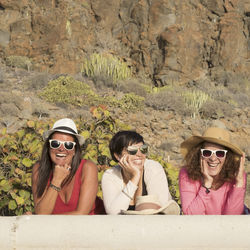 Smiling friends wearing sunglasses standing by retaining wall against mountain