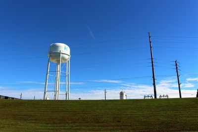 Water tower by telephone poles on field against blue sky