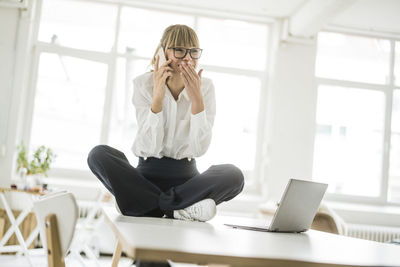 Happy businesswoman sitting on desk in office talking on cell phone