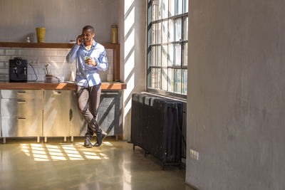 Young entrepreneur standing in company kitchen, drinking coffee, using smartphone