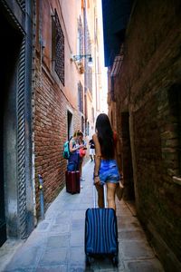 Rear view of young woman with luggage walking on alley amidst buildings in city