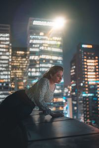 Side view of man sitting against illuminated buildings in city at night