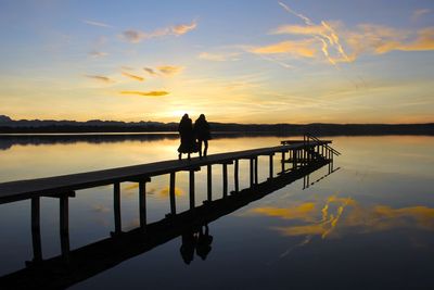 Silhouette people walking on pier over lake against sky during sunset