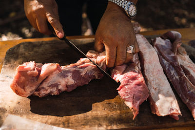 Crop unrecognizable man with watches and ring on finger butchering meat with knife on wooden cutting board preparing food outdoors