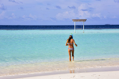 Rear view of woman wearing bikini walking at sea shore against sky