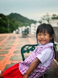 Portrait of cute smiling girl sitting on bench against sky