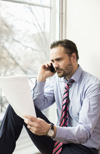 Serious mid adult businessman reading document while on call at office
