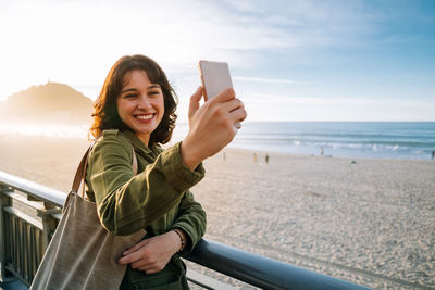 Portrait of smiling young man using mobile phone at beach against sky