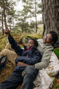 Boy pointing while lying down with father near tree in forest