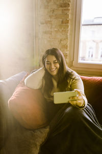 Happy woman taking selfie through mobile phone sitting on sofa at home