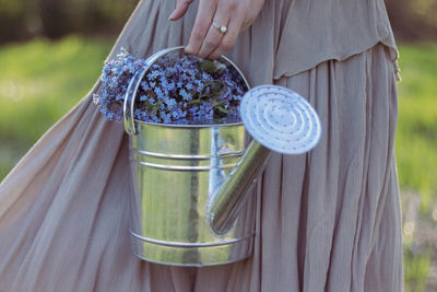 Midsection of a woman holding a watering can full of small blue and purple flowers.