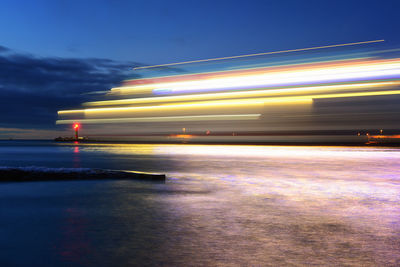 Light trails on city by sea against sky at night