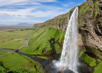 Scenic view of majestic waterfall and small rainbow against expansive landscape sky