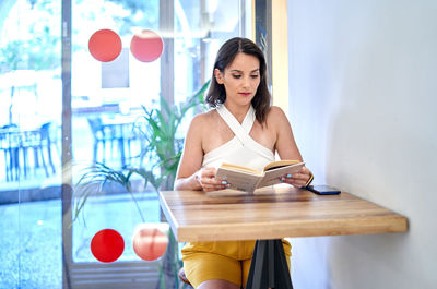 Attentive young female reading interesting story in book while sitting at table and spending weekend in cafe