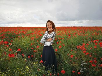 Portrait of woman standing at red poppy farm against cloudy sky