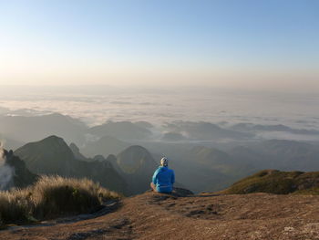 Rear view of man sitting on mountain white looking at foggy landscape