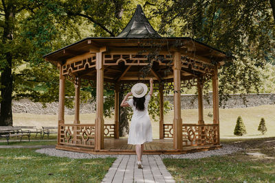 Rear view of beautiful young woman wearing white dress standing under wooden pavilion in park