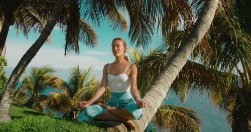 Girl meditates on grassy lawn in a lotus position and breathes deeply. tropical palm trees on ocean.