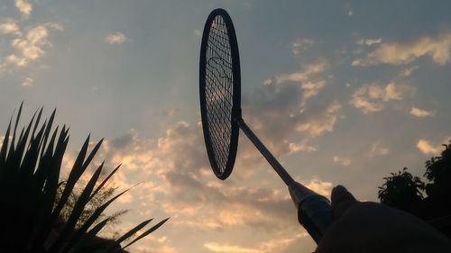 Cropped image of hand holding badminton racket against cloudy sky during sunset