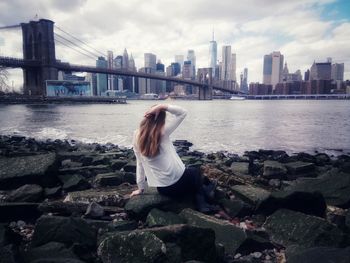 Rear view of woman looking at modern buildings while sitting on rock by river