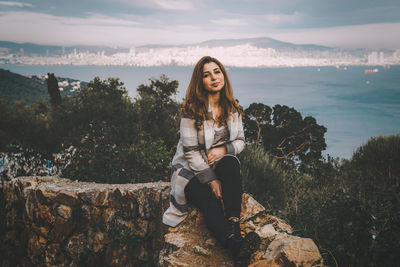 Portrait of young woman sitting on rock against sky