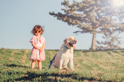 Cute stylish little caucasian child girl in sunglasses walking dog in park field on summer day