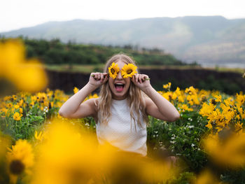 Excited woman shouting while holding flowers over eyes by plants against sky