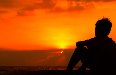 Silhouette man sitting on beach against orange sky