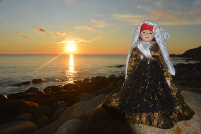 Woman on rock at beach against sky during sunset