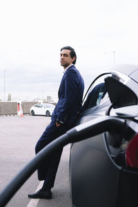 Businessman with hand in pocket leaning on car charging at electric station